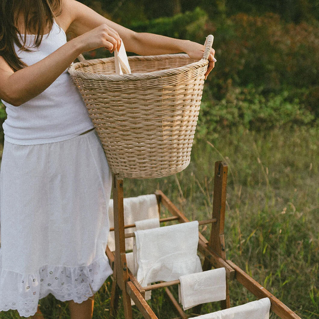 Woman carrying a woven basket of linens beside a drying rack in a garden